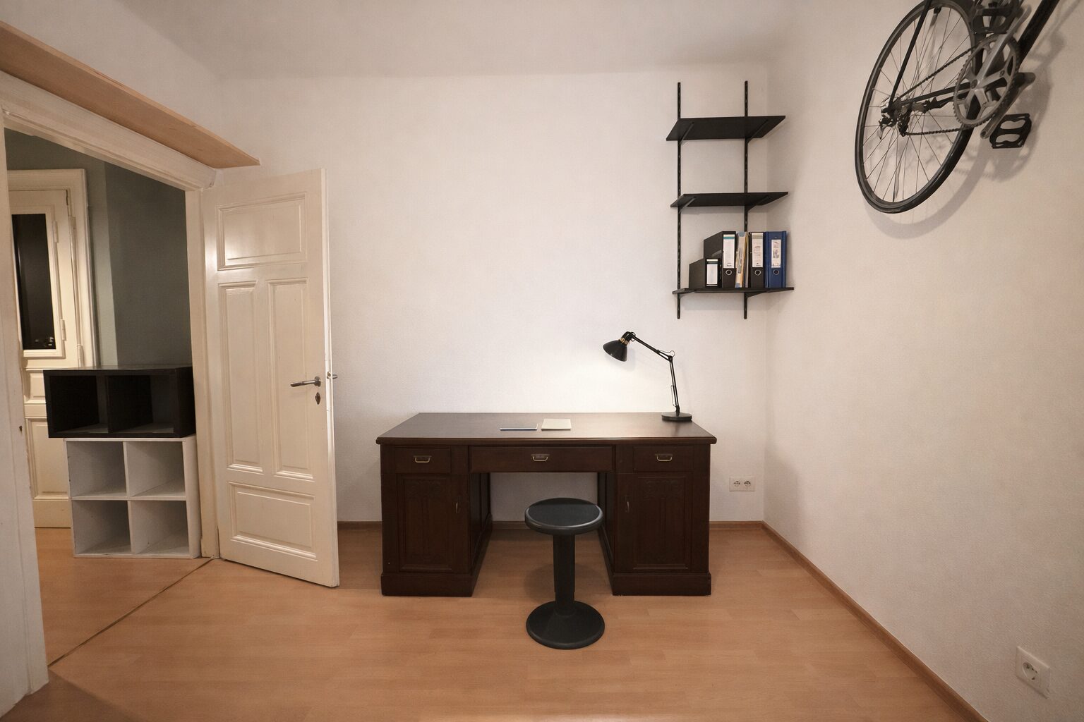 Home office with a dark wooden desk, black task lamp, and a round stool; wall-mounted shelves hold binders. A bicycle is hung on the right wall.