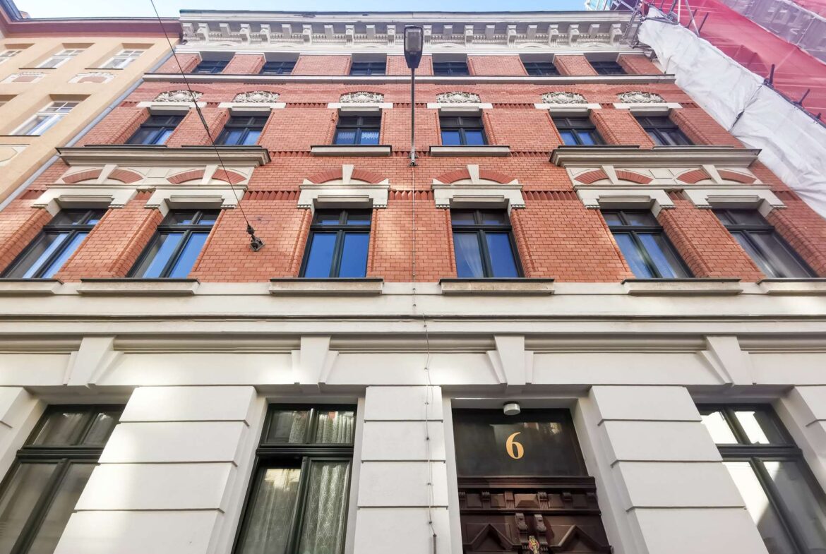 Red brick building facade with white decorative trim, tall windows, and a gold-numbered black door (6) at street level, centered under a streetlamp.