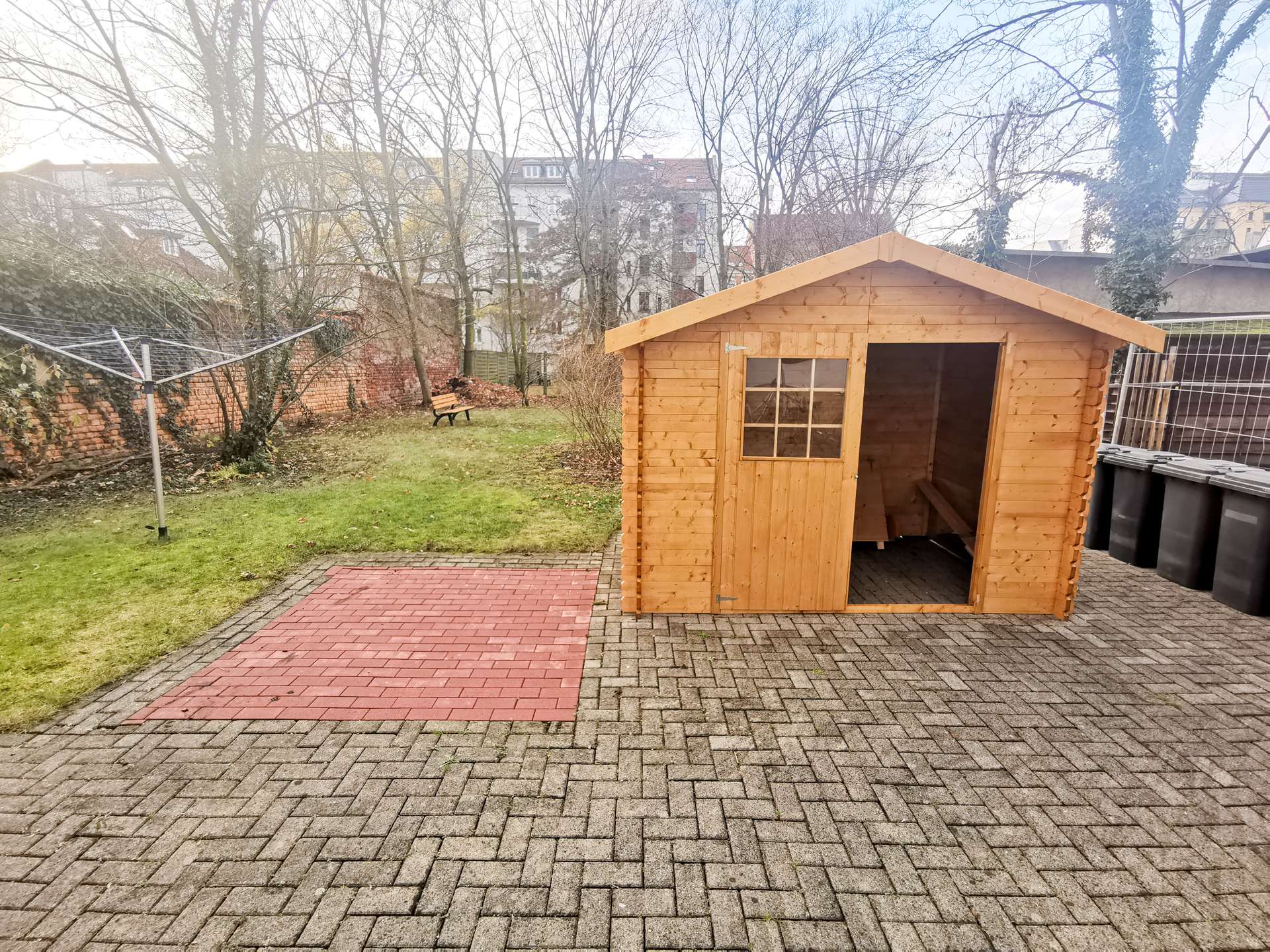 Backyard scene with a wooden garden shed on the right, brick and paver patio, and leafless trees in the background.