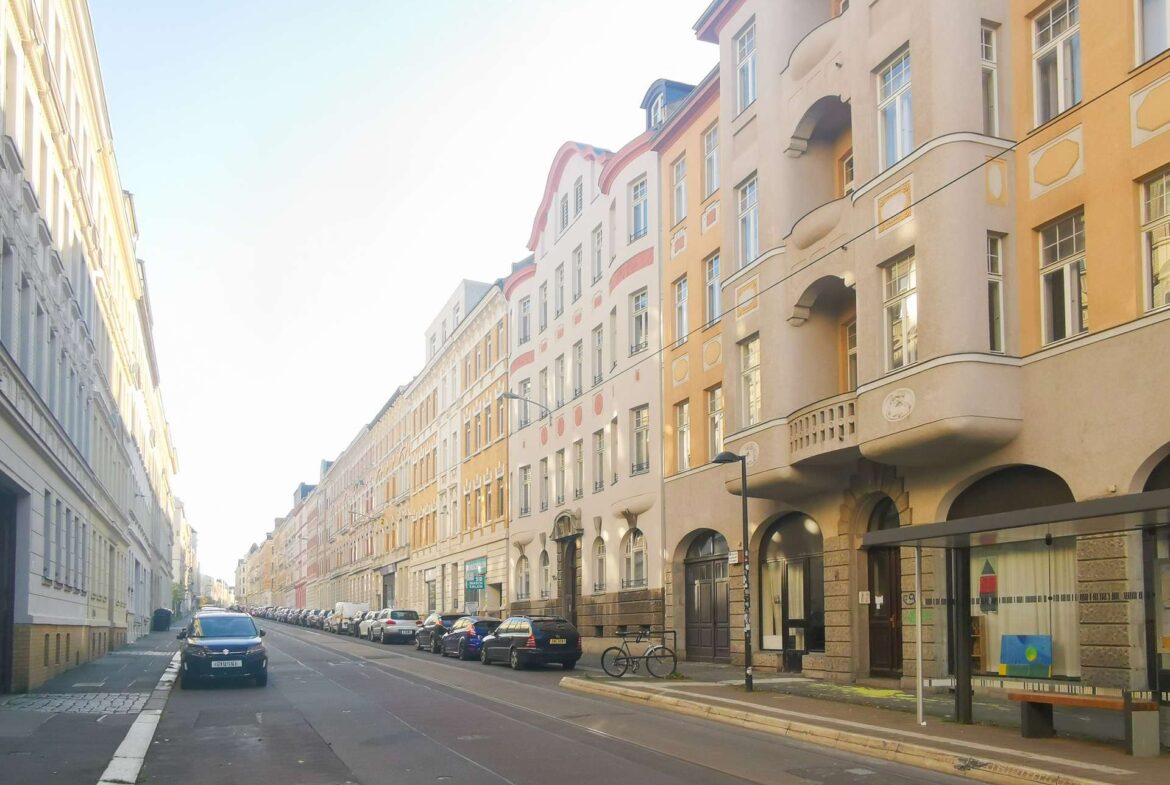 City street lined with pastel-colored apartment buildings, parked cars, and tram tracks along the road on a sunny day