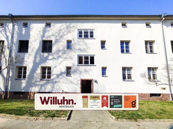 White multi-story residential building with a large Willuhn Immobilien sign in front and awards plaques nearby.