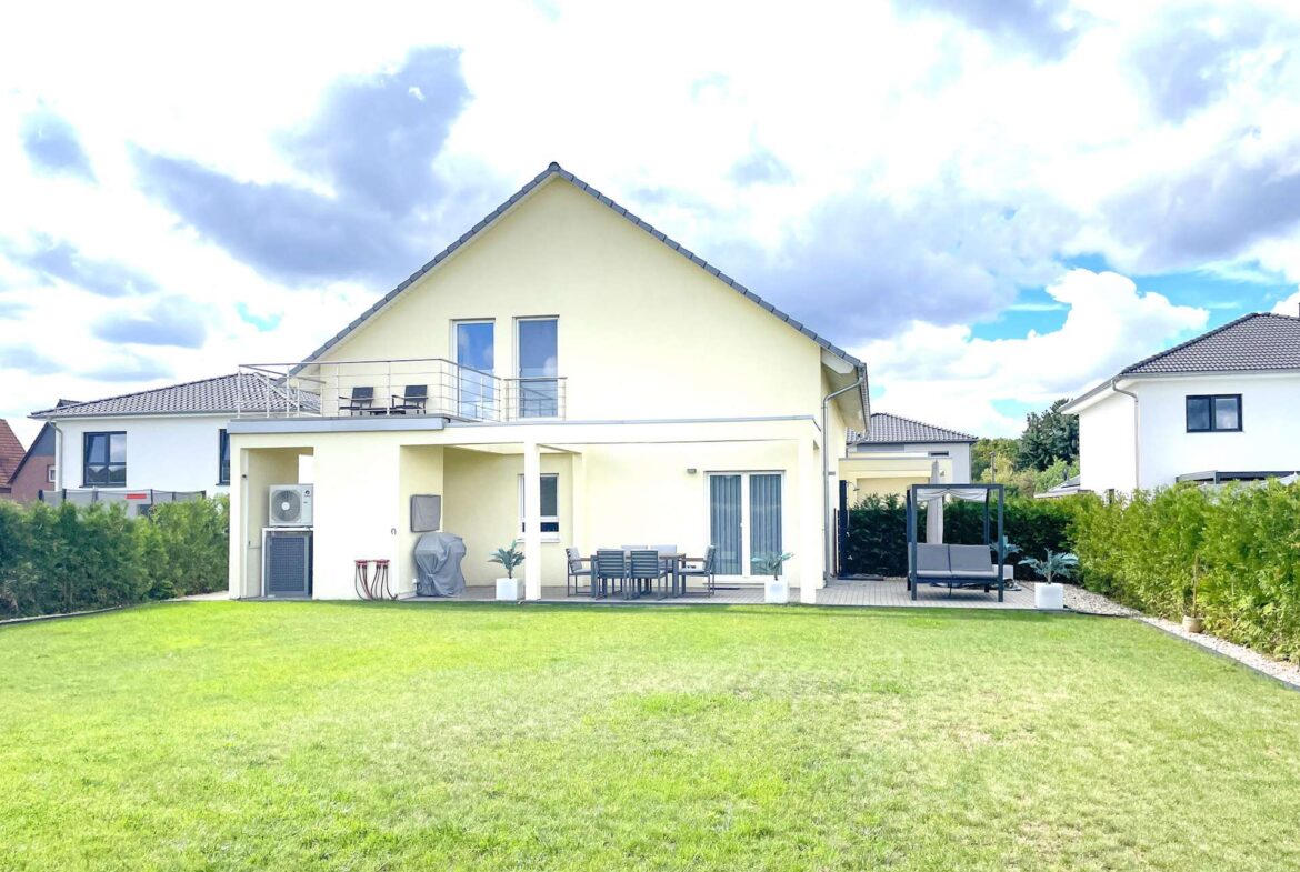 Backyard of a light-yellow two-story house with a large green lawn, a covered patio with outdoor dining table, chairs, and a grill area, plus a hedge along the fence and a gazebo on a paved section to the right.
