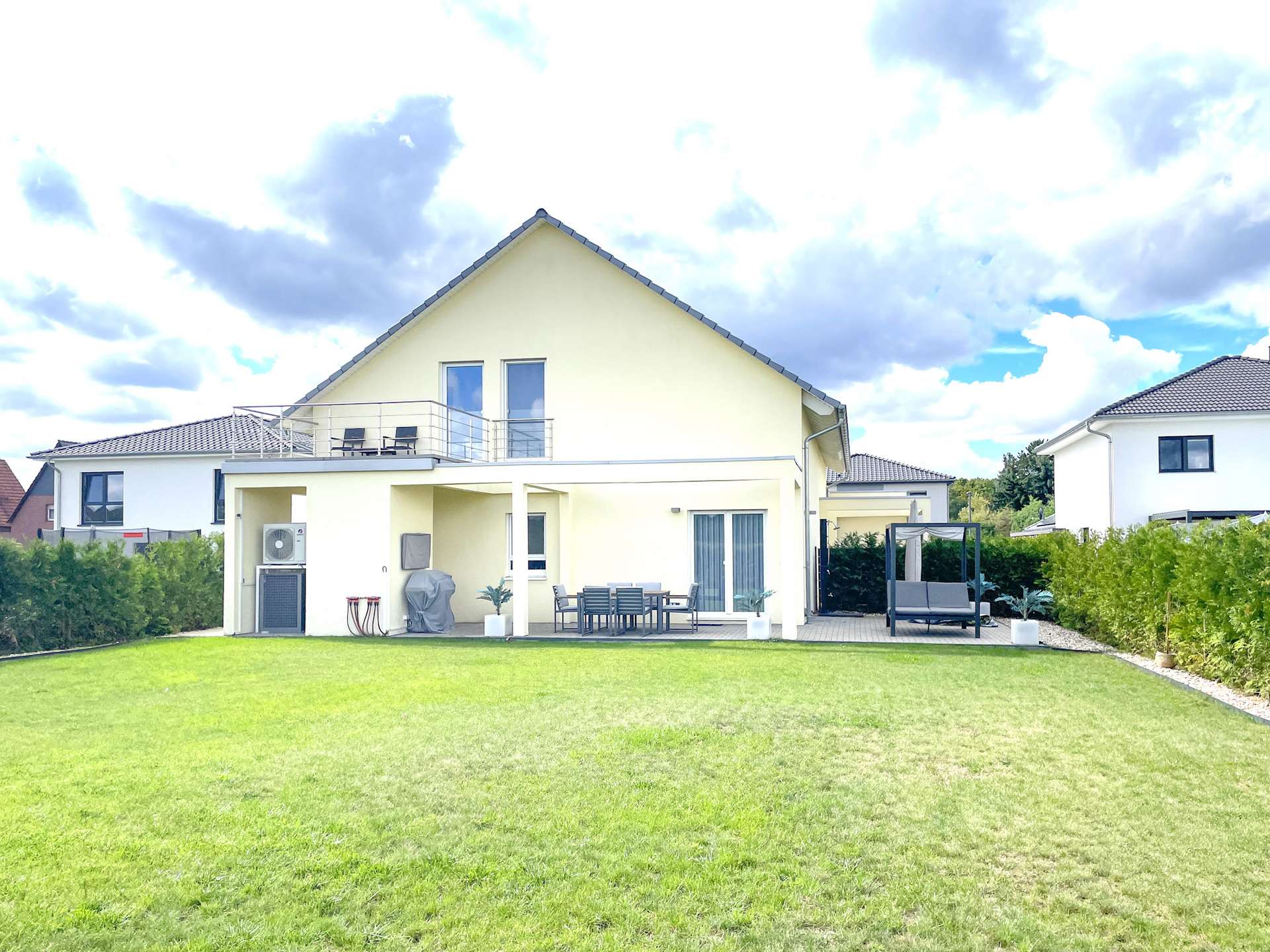 Backyard of a light-yellow two-story house with a large green lawn, a covered patio with outdoor dining table, chairs, and a grill area, plus a hedge along the fence and a gazebo on a paved section to the right.