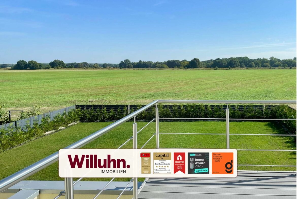 Balcony railing with a Willuhn Immobilien sign and award stickers, overlooking a green field and distant trees under a bright blue sky.