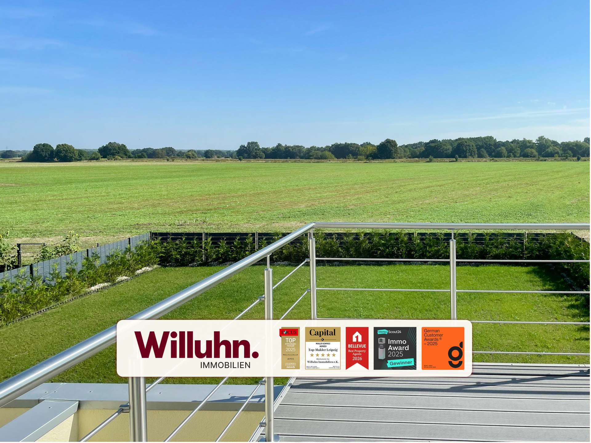 Balcony railing with a Willuhn Immobilien sign and award stickers, overlooking a green field and distant trees under a bright blue sky.
