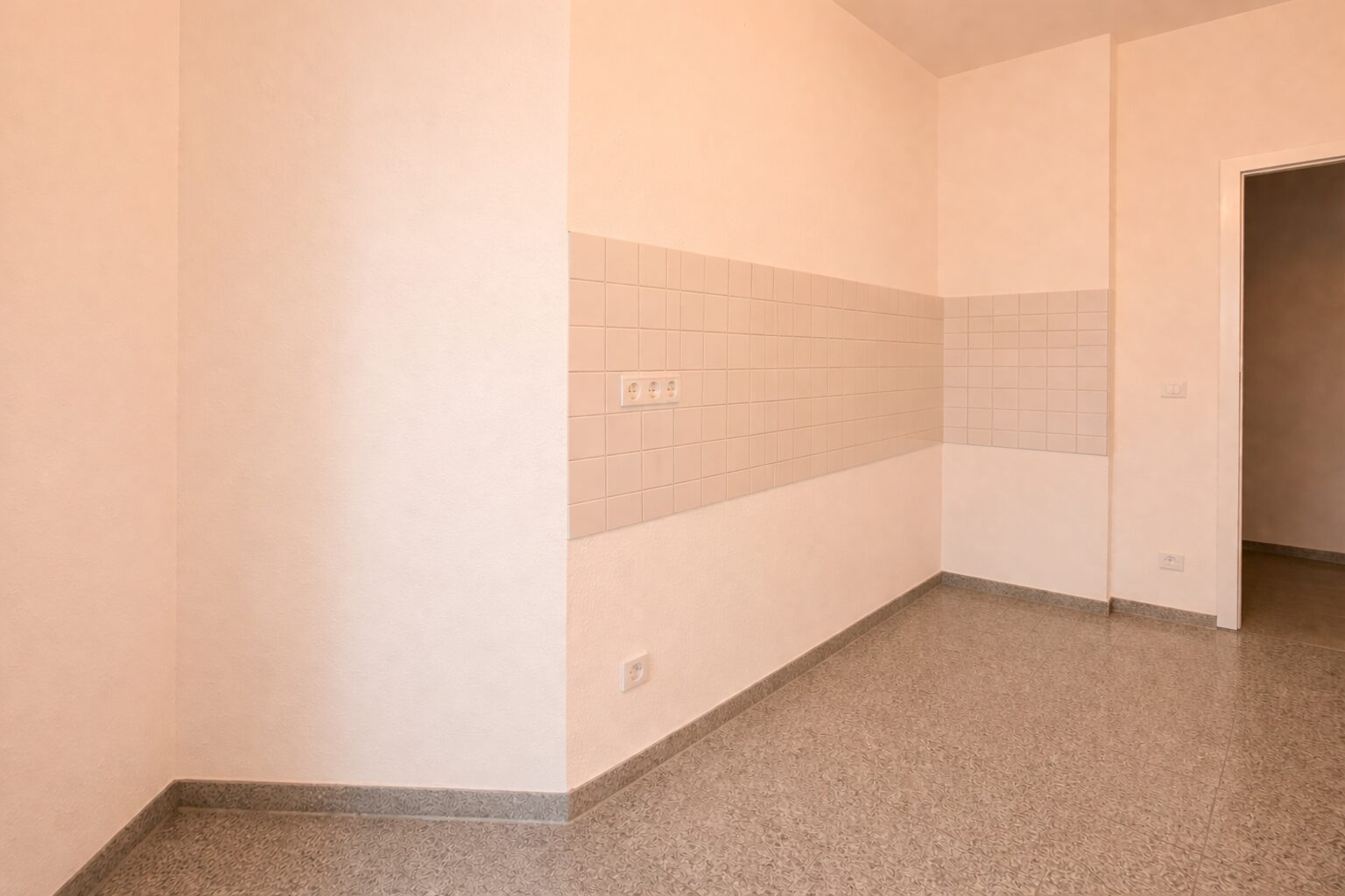 Empty room with beige walls and a horizontal tiled panel along the middle of the wall, featuring three electrical outlets and a speckled floor.