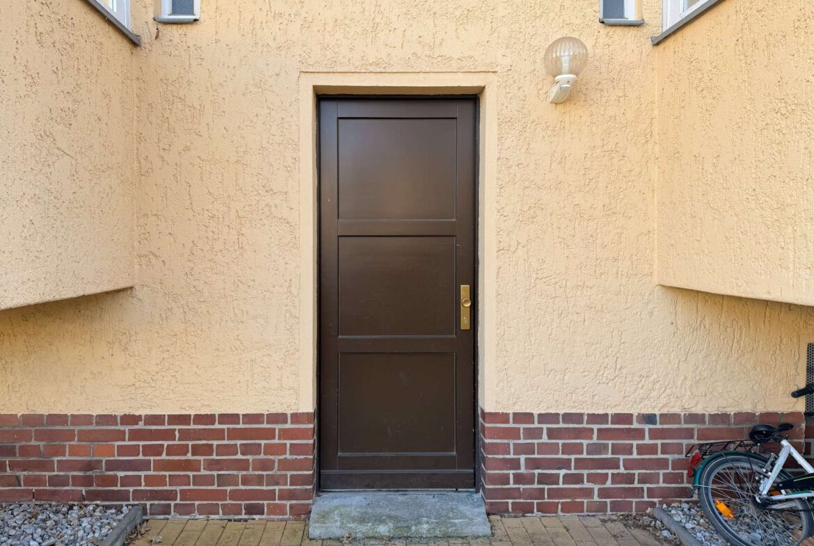Brown paneled entrance door set in a beige stucco wall with a brick base and a round exterior light above, bicycle leaned nearby on the right.