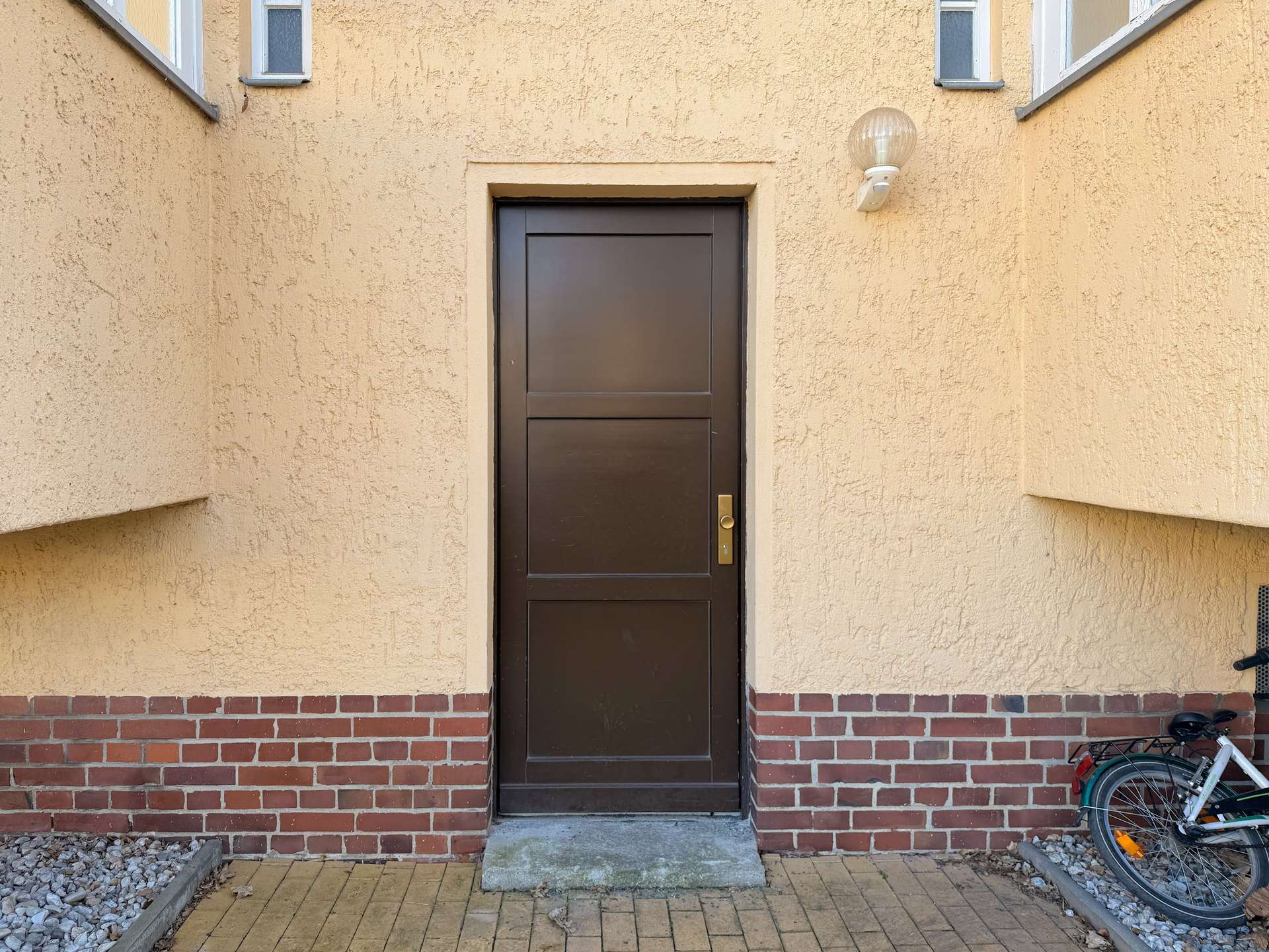 Brown paneled entrance door set in a beige stucco wall with a brick base and a round exterior light above, bicycle leaned nearby on the right.
