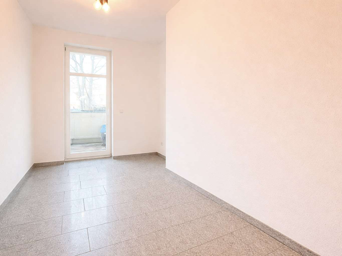 Empty small room with pale walls, gray tiled floor, and a glass door opening to a balcony with a tree outside.