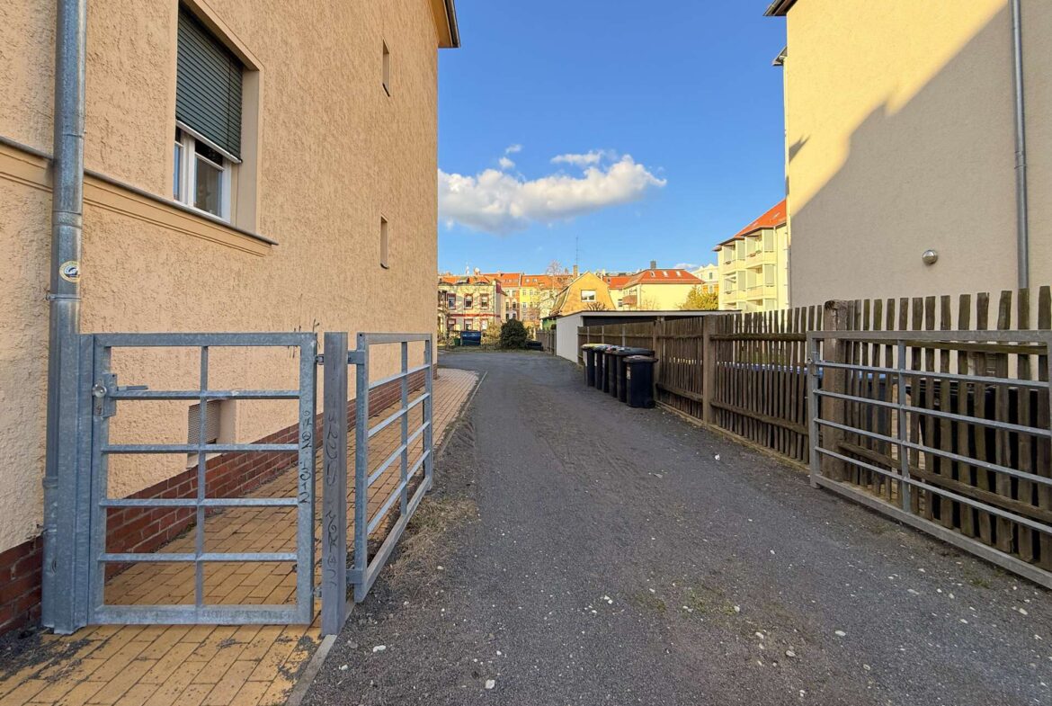 Narrow alley between a beige apartment building on the left and a wooden fence on the right, with a metal gate and trash bins along the path under a blue sky.