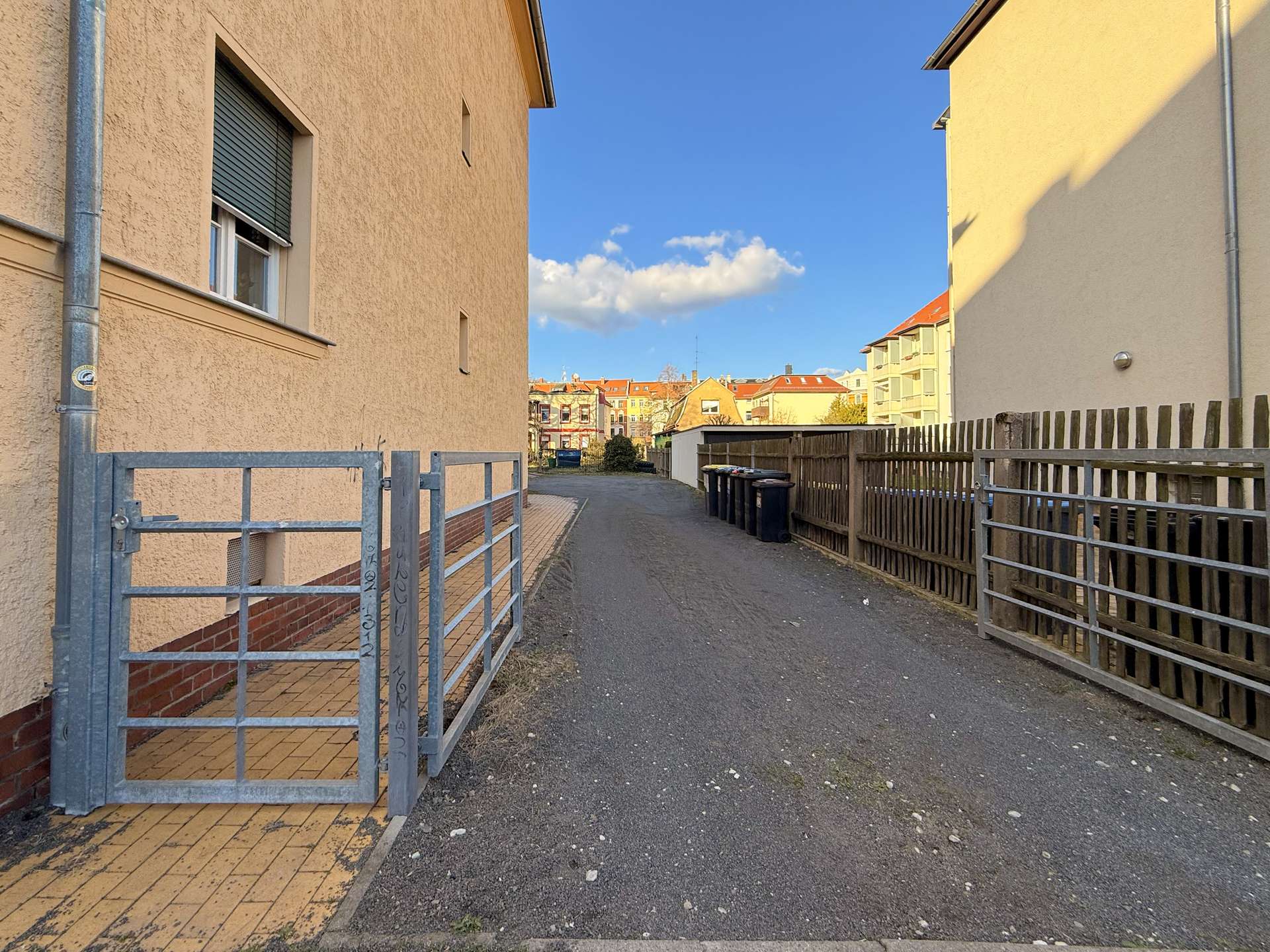 Narrow alley between a beige apartment building on the left and a wooden fence on the right, with a metal gate and trash bins along the path under a blue sky.