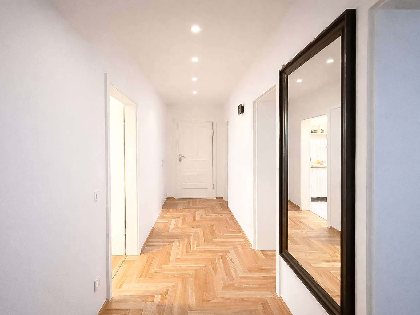 Bright white hallway with a herringbone wood floor, recessed ceiling lights, and a large dark-framed mirror on the right.