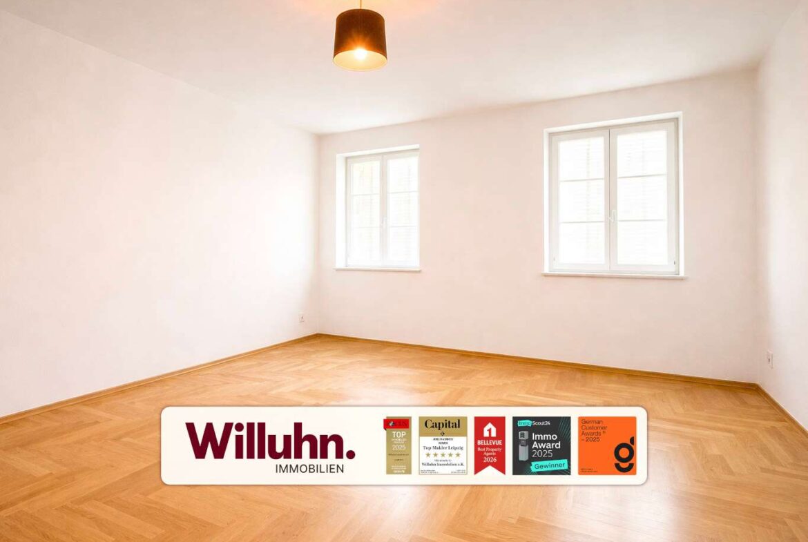 Bright empty room with parquet floor, two windows, and a ceiling light; real estate branding on the floor.