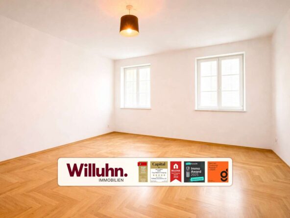Bright empty room with parquet floor, two windows, and a ceiling light; real estate branding on the floor.
