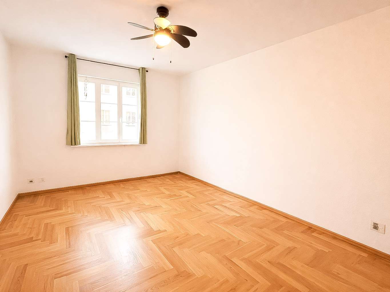 Empty room with light wood herringbone floor, white walls, a window with green curtains, and a ceiling fan with light on.