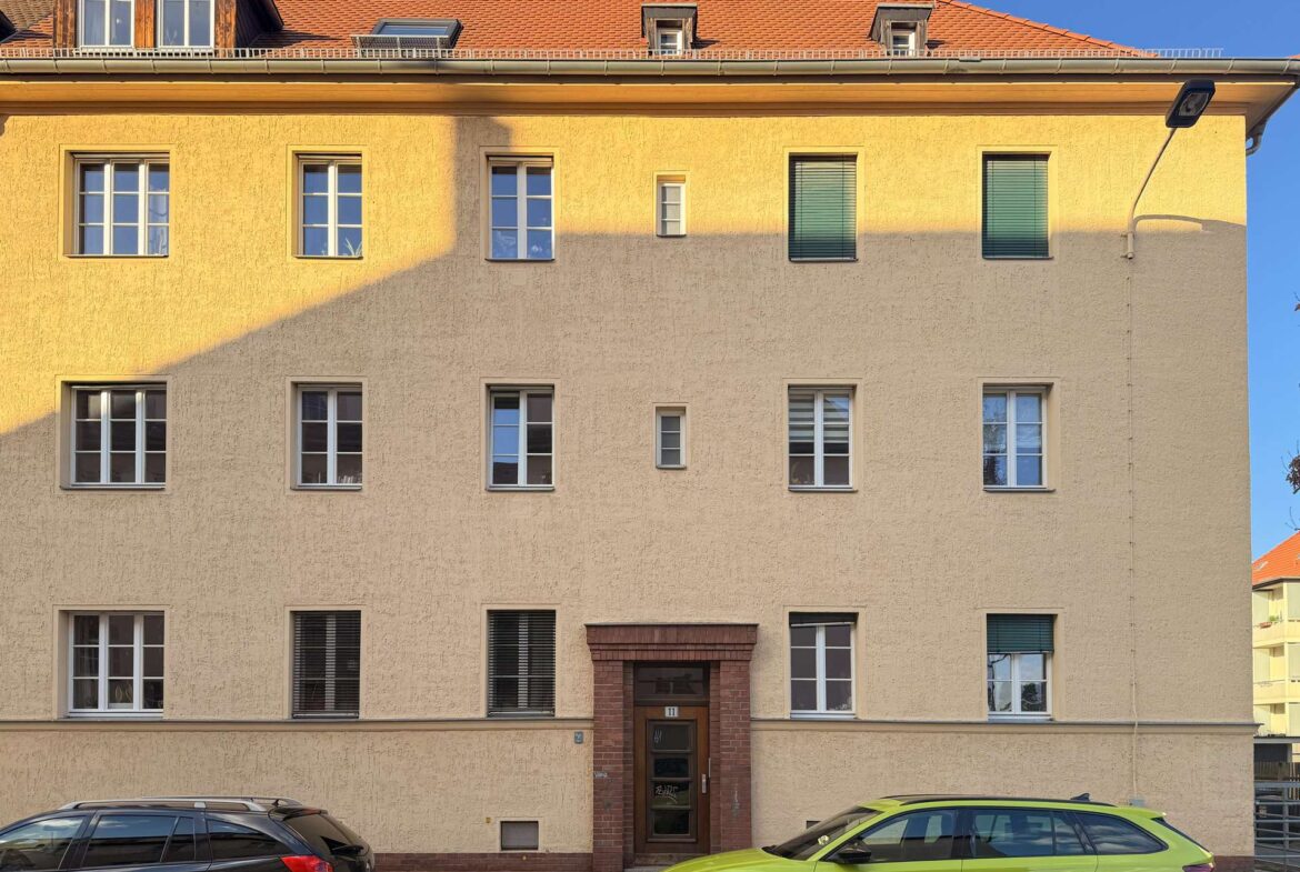 Facade of a beige apartment building with a red-tiled roof, multiple white-framed windows, a brick entryway, and two cars parked in front (black and lime-green).