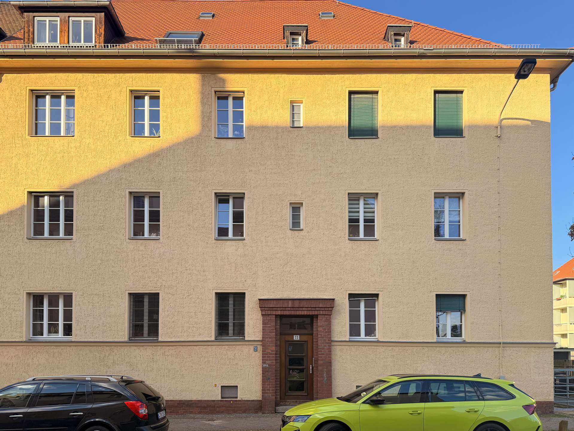 Facade of a beige apartment building with a red-tiled roof, multiple white-framed windows, a brick entryway, and two cars parked in front (black and lime-green).