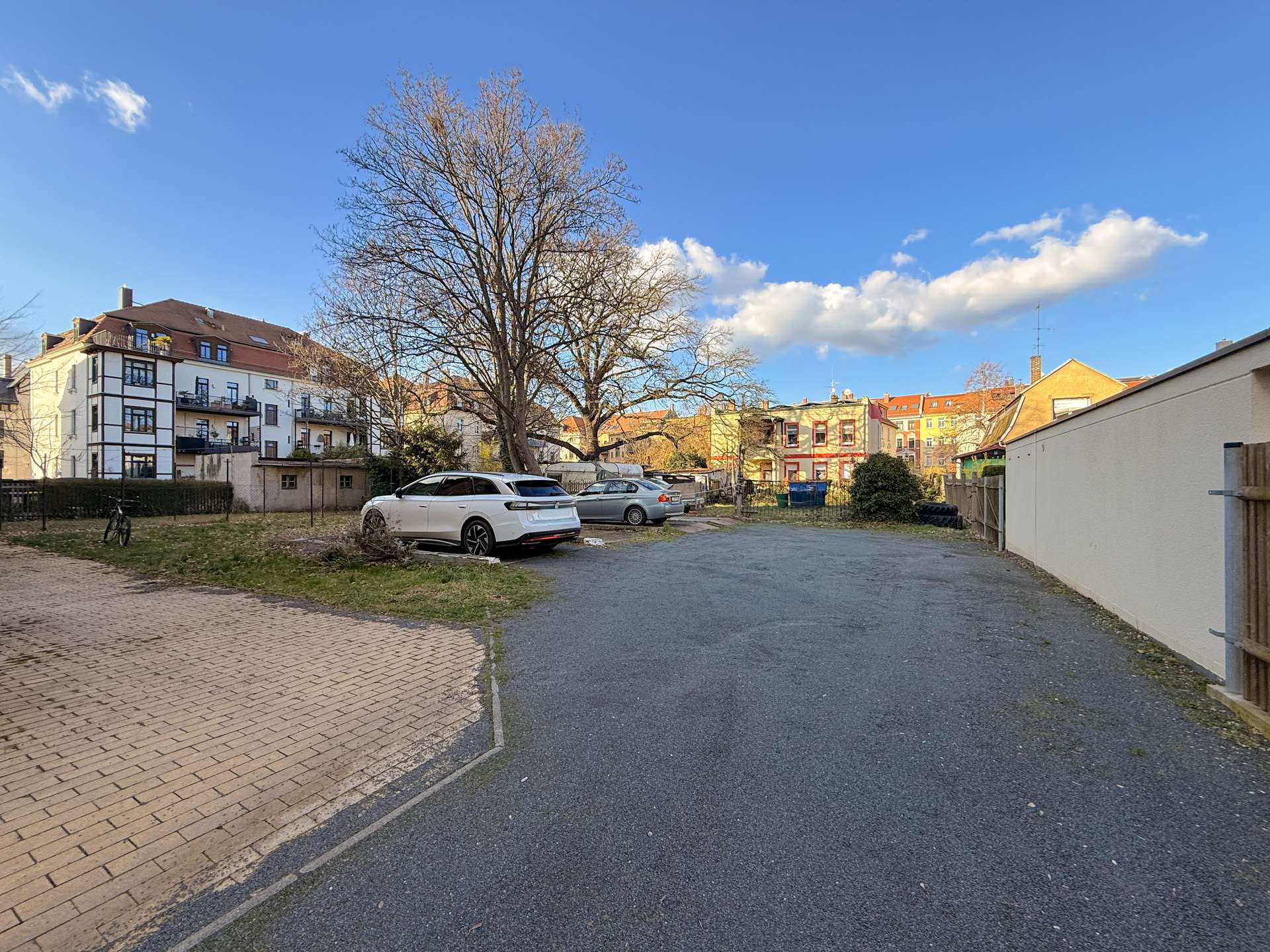 Residential parking lot with several cars, a large leafless tree, and apartment buildings under a blue sky in a quiet neighborhood