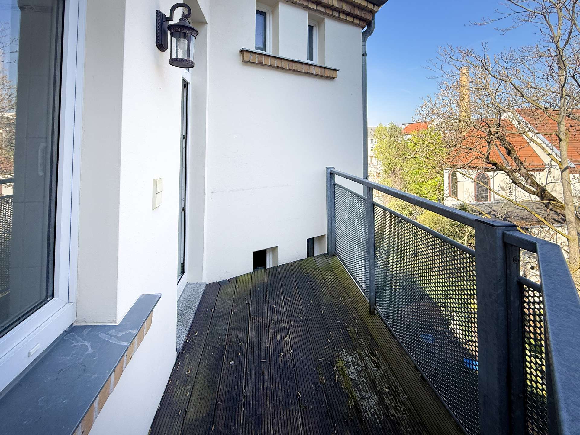 Narrow wooden balcony walkway along a white building with a metal mesh railing, blue sky and leafless trees in the background.
