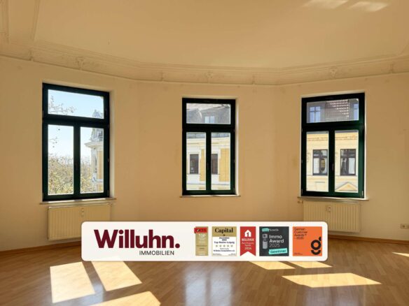 Sunlit empty room with three green-framed windows and a curved white Willuhn Immobilien banner with award logos at the bottom panel.