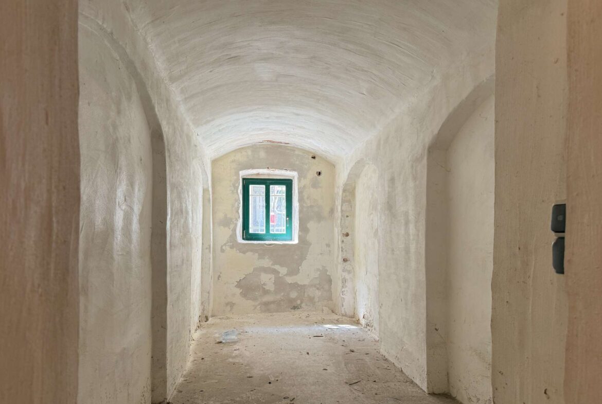 Narrow corridor with arched white ceiling and peeling beige walls, ending in a green-framed window at the far end.