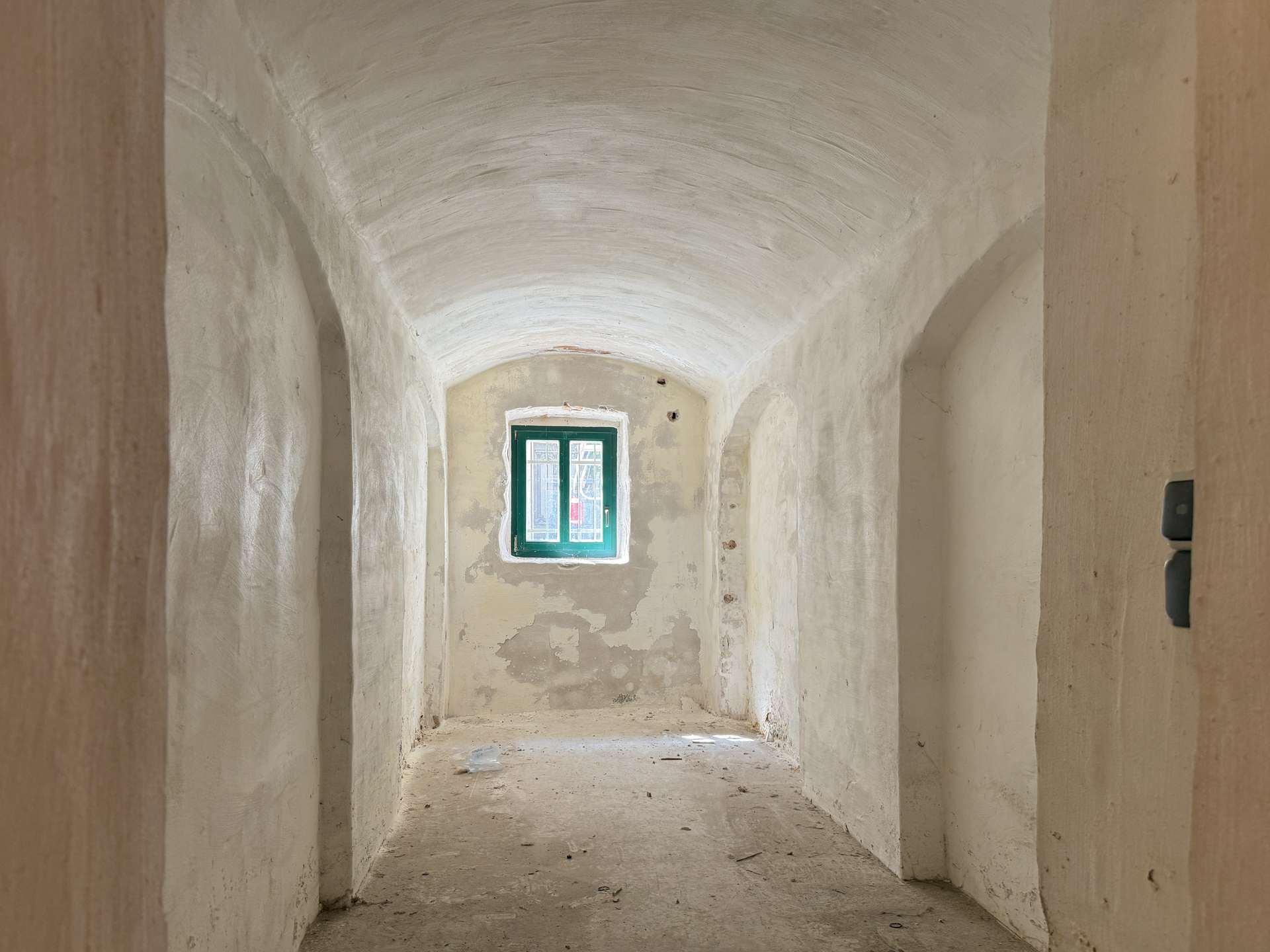 Narrow corridor with arched white ceiling and peeling beige walls, ending in a green-framed window at the far end.
