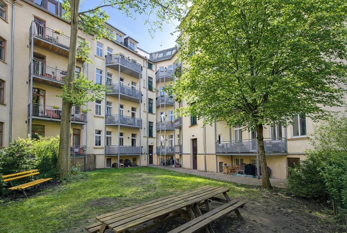 Residential courtyard with yellow apartment building, multiple balconies, and leafy trees; a wooden picnic table and bench in the foreground.