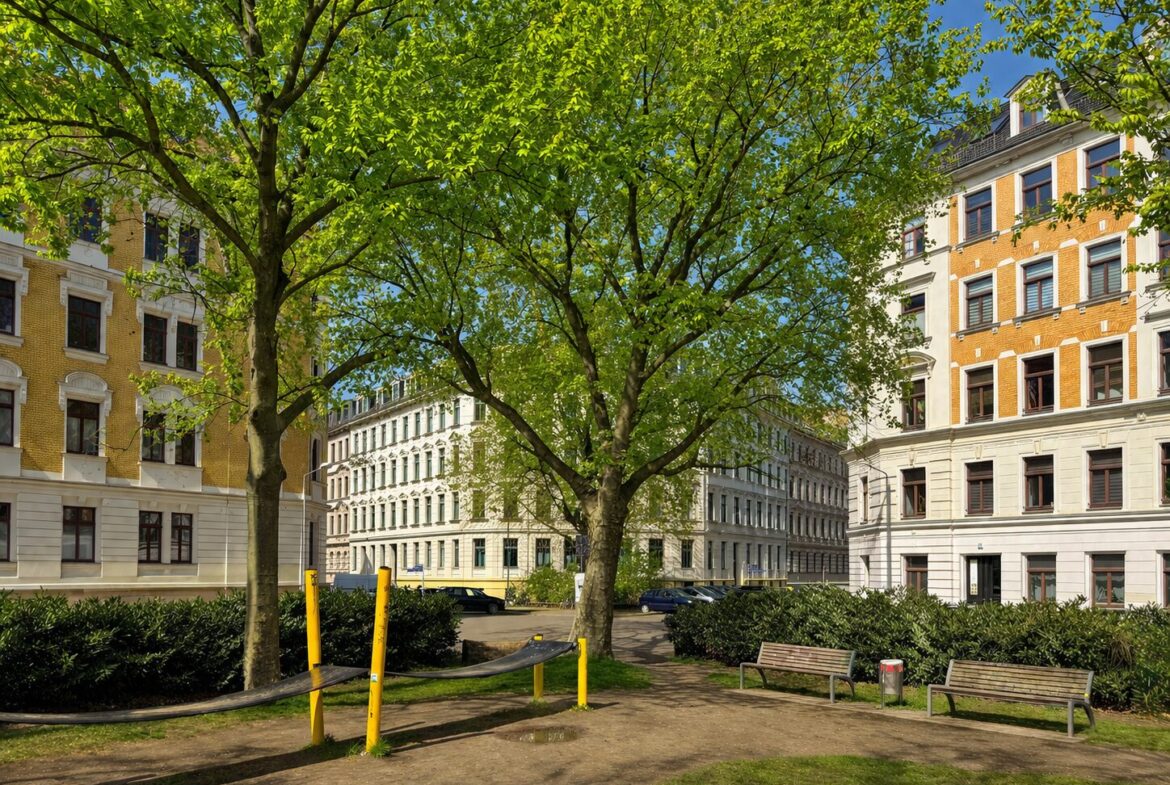 City park with large green trees, benches, and light-colored apartment buildings surrounding the yard.