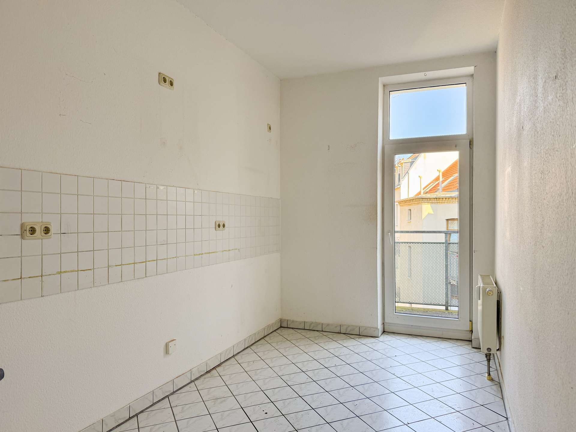 Empty white kitchen/utility corner with tiled wall, tile floor, and a glass door to a balcony showing buildings outside; radiator on right side.