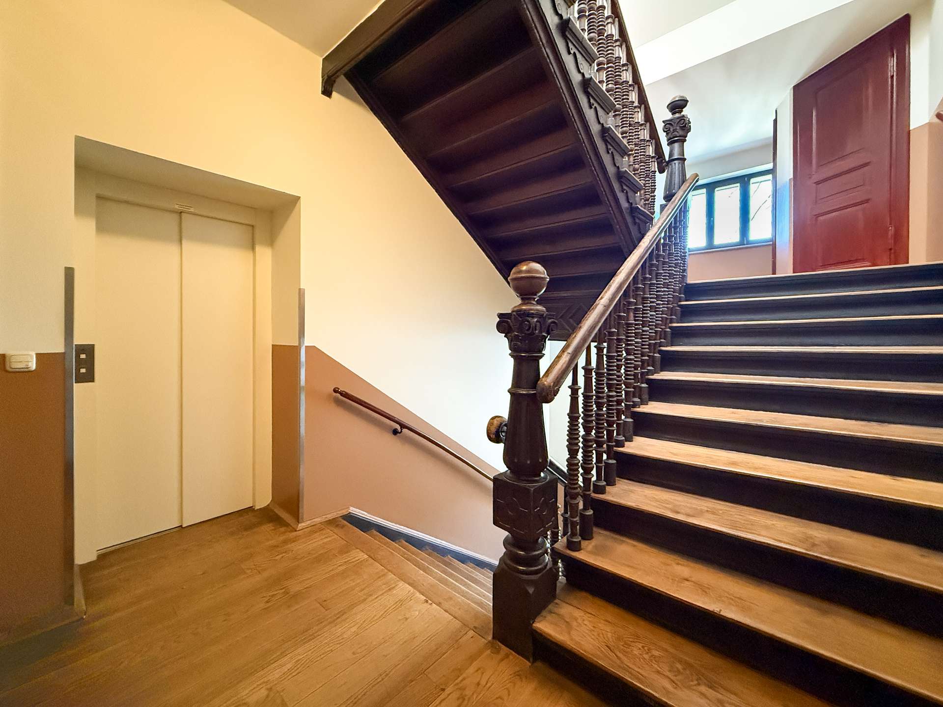 Interior stairwell with a dark wooden staircase and ornate balusters, beige walls, and an elevator on the left side.