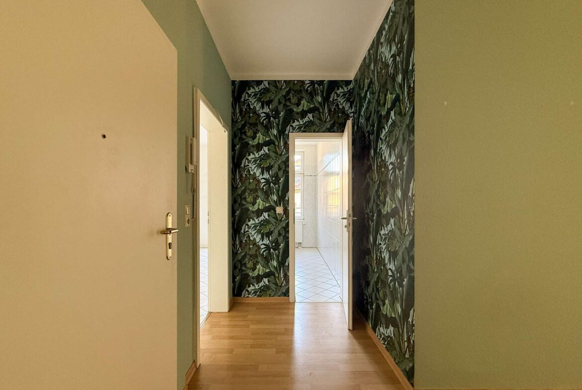 Narrow hallway with pale green walls and bold jungle-leaf wallpaper on the right, leading to a sunlit, white-tiled room at the end.