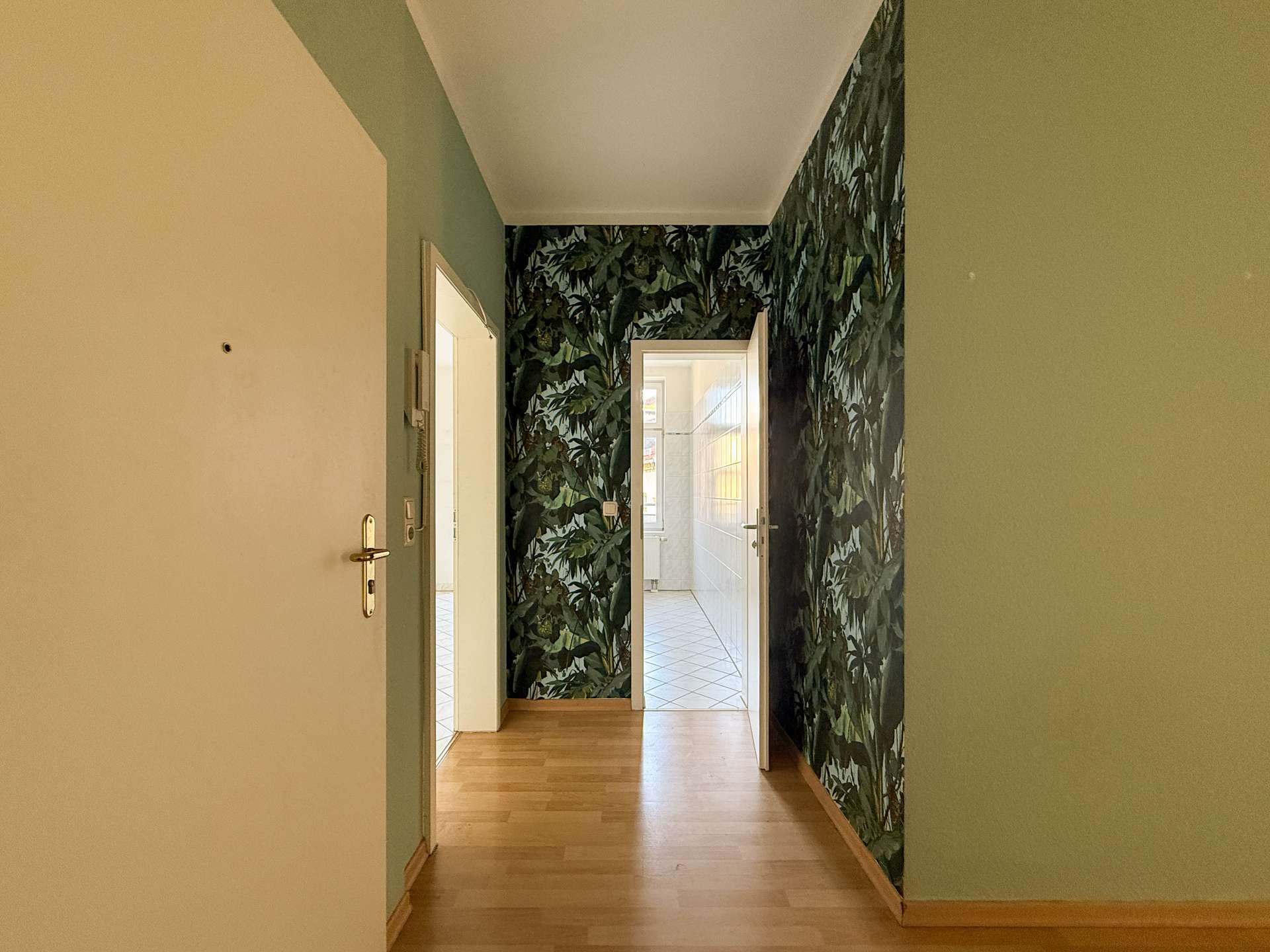 Narrow hallway with pale green walls and bold jungle-leaf wallpaper on the right, leading to a sunlit, white-tiled room at the end.