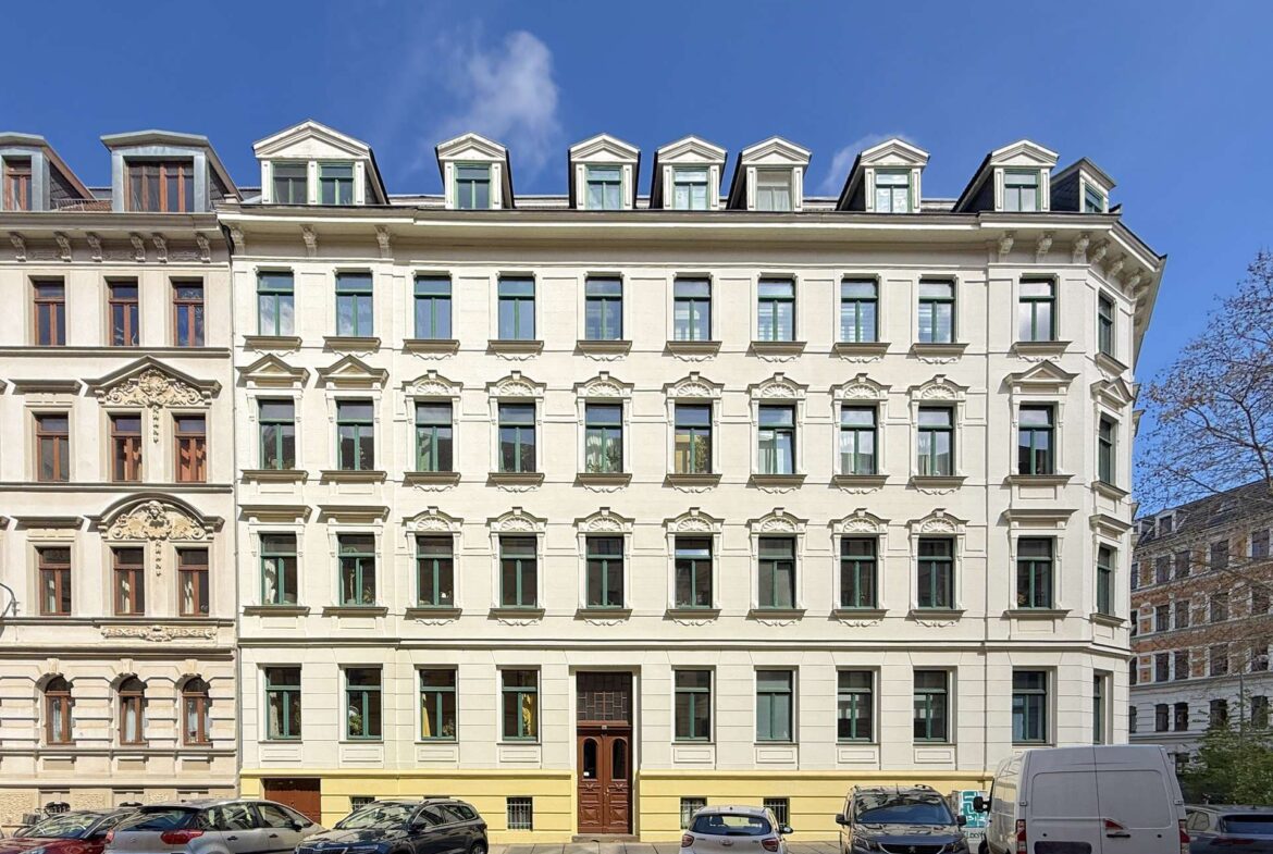 Facade of a pale historic building with decorative moldings, rows of green-trimmed windows, and parked cars in front under a blue sky.