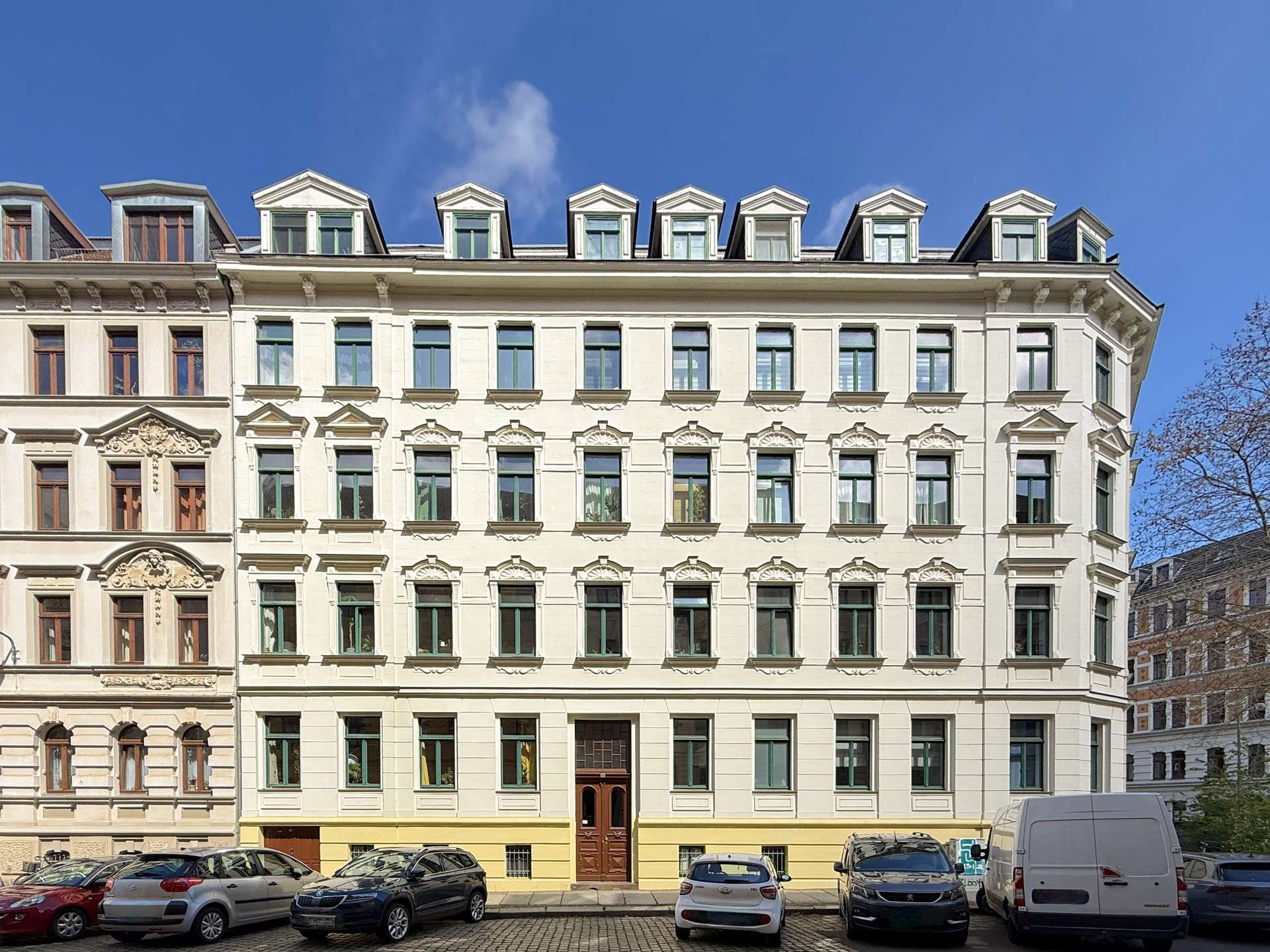 Facade of a pale historic building with decorative moldings, rows of green-trimmed windows, and parked cars in front under a blue sky.
