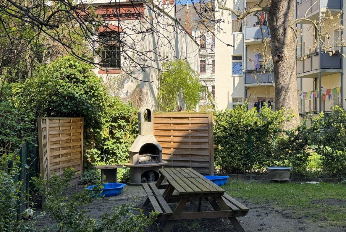 Backyard with a concrete grill and wooden picnic table amid green plants and a wooden fence, apartment building in the background.