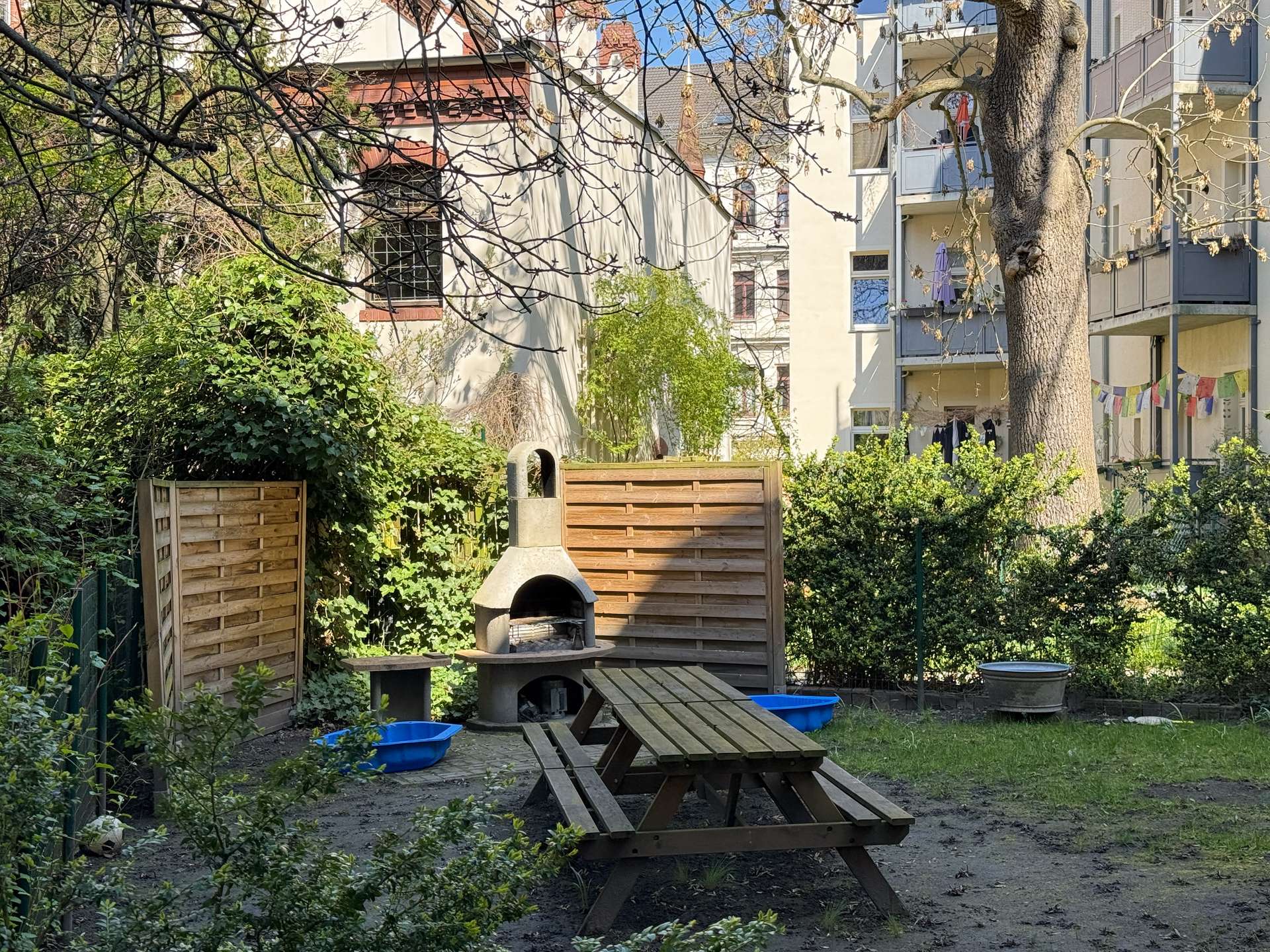 Backyard with a concrete grill and wooden picnic table amid green plants and a wooden fence, apartment building in the background.