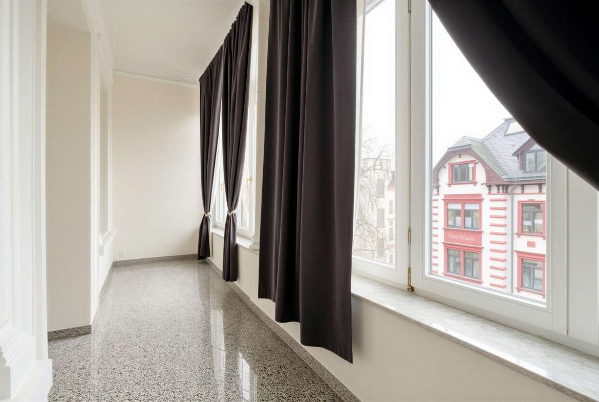Bright indoor corridor with large windows and dark floor-length curtains, overlooking a red-and-cream building outside.