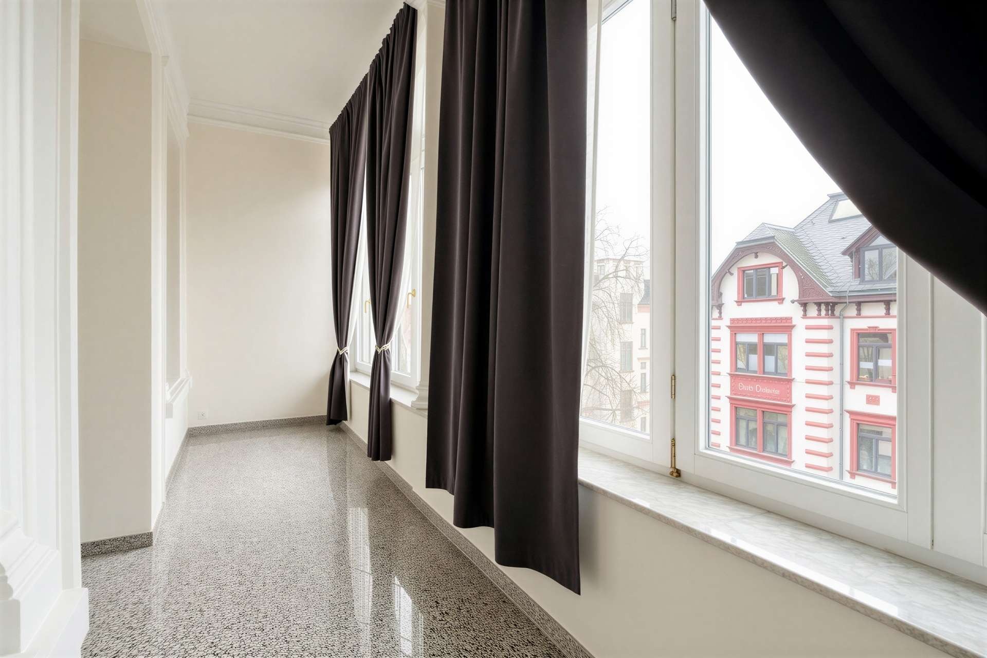 Bright indoor corridor with large windows and dark floor-length curtains, overlooking a red-and-cream building outside.