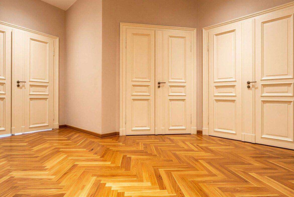 Hallway with cream paneled doors along beige walls and a herringbone wood floor, warmly lit.