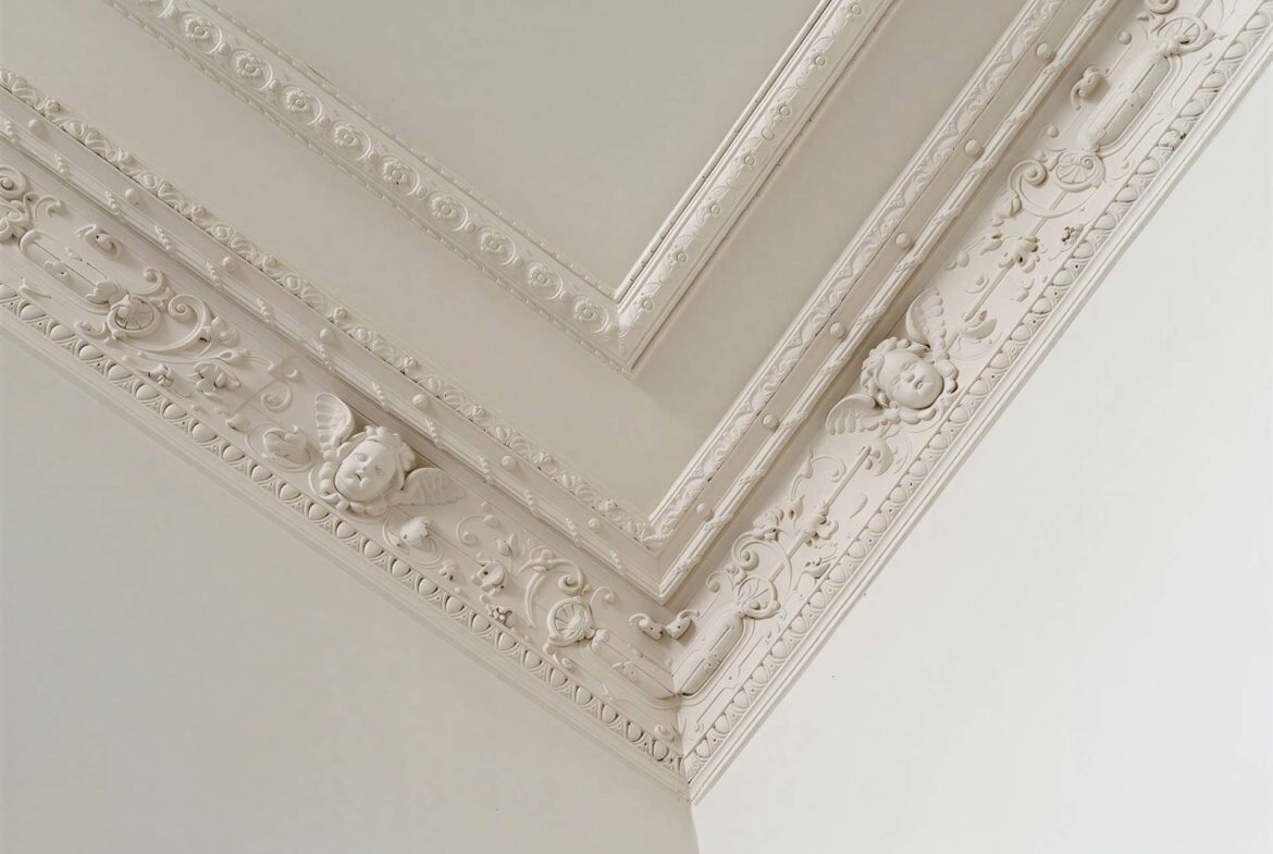 Ornate white plaster molding along a ceiling corner with floral scrolls and cherub faces.