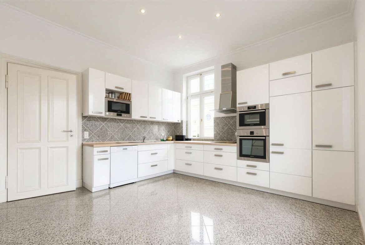 Bright white kitchen with glossy cabinets, stainless steel double ovens, and a gray tiled backsplash beneath a window.