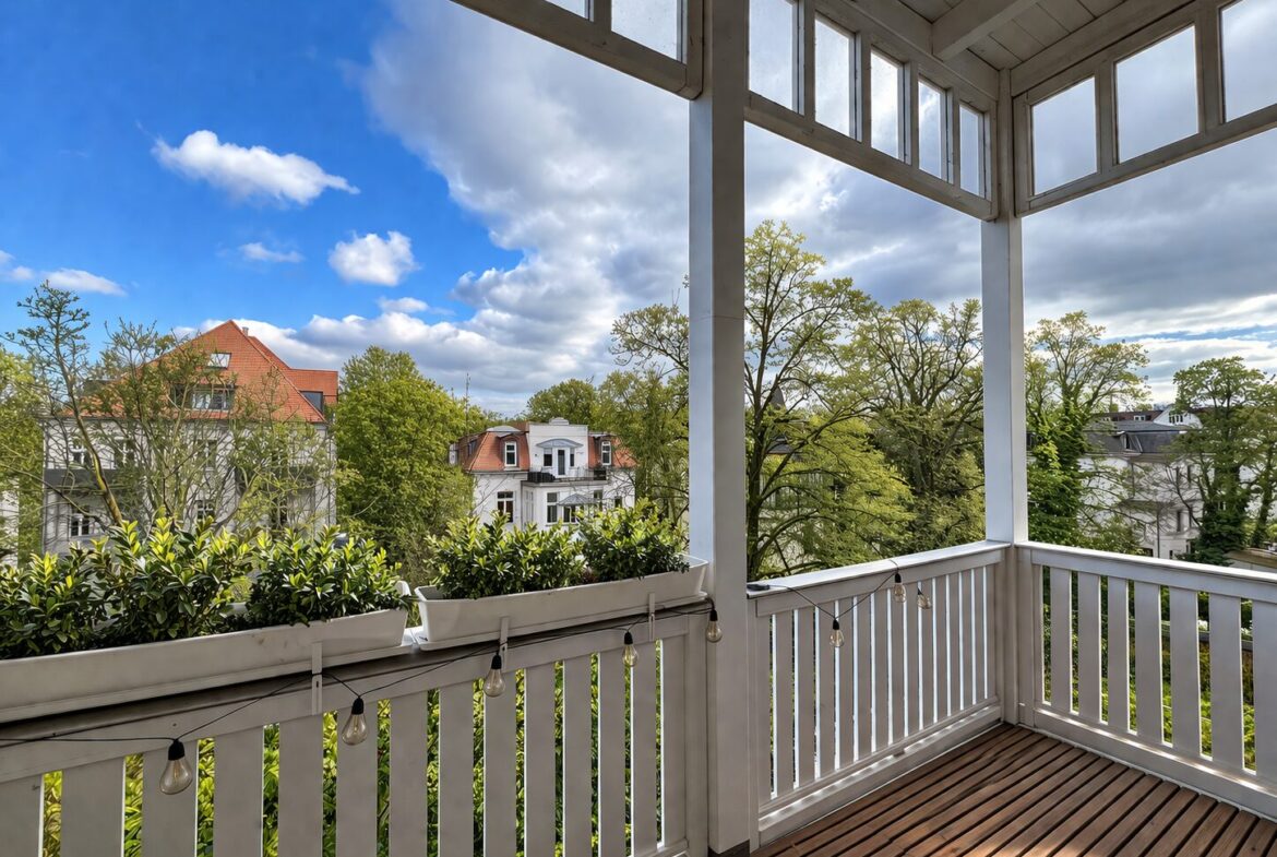 Balcony with white railing and string lights; overlooks trees and white houses under a blue sky, sunny day.