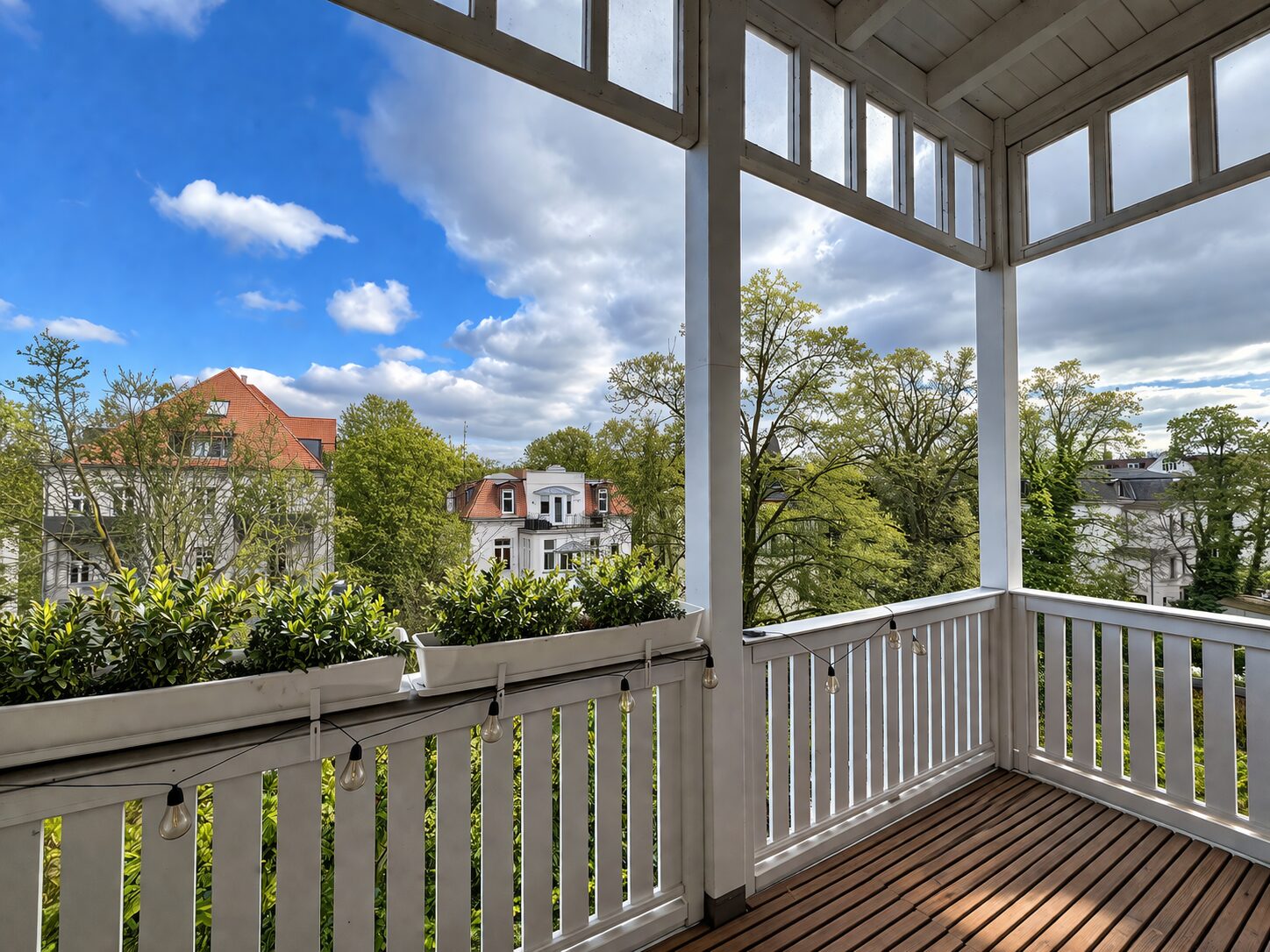 Balcony with white railing and string lights; overlooks trees and white houses under a blue sky, sunny day.