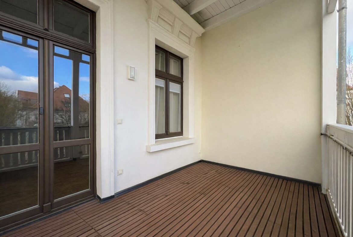 Balcony with brown-framed glass doors, a window, white walls, and a wooden plank deck with a railing along the edge.