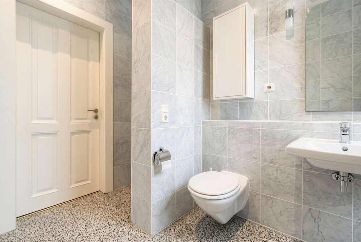 Bright marble-tiled bathroom with a white door, wall cabinet, toilet, and wall-mounted sink.