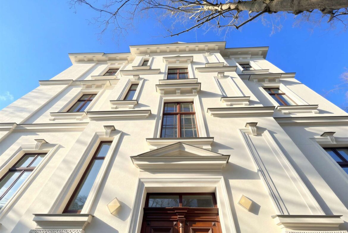 White neoclassical apartment building viewed from street level against a clear blue sky, with bare tree branches overhead.