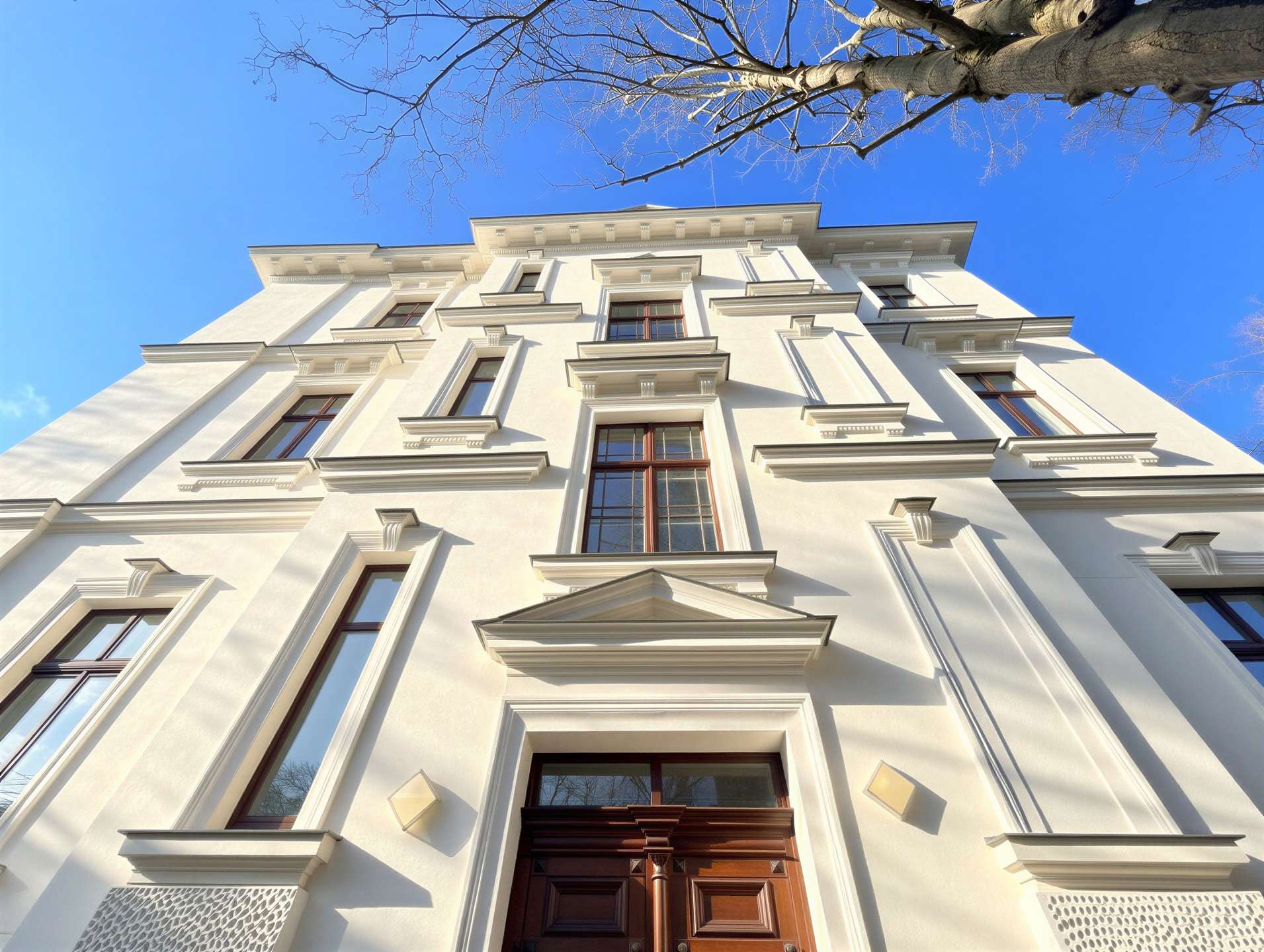 White neoclassical apartment building viewed from street level against a clear blue sky, with bare tree branches overhead.