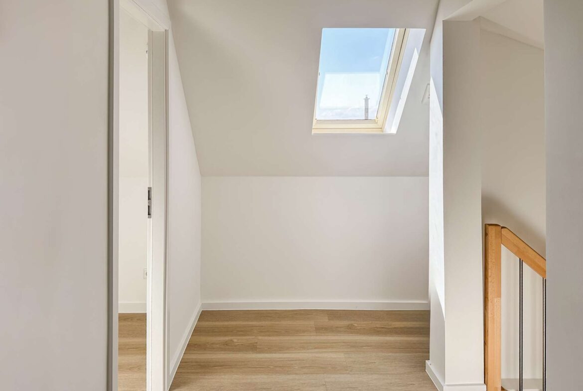 Bright upstairs hallway with an angled ceiling and a large skylight letting in daylight, white walls and a wooden handrail.