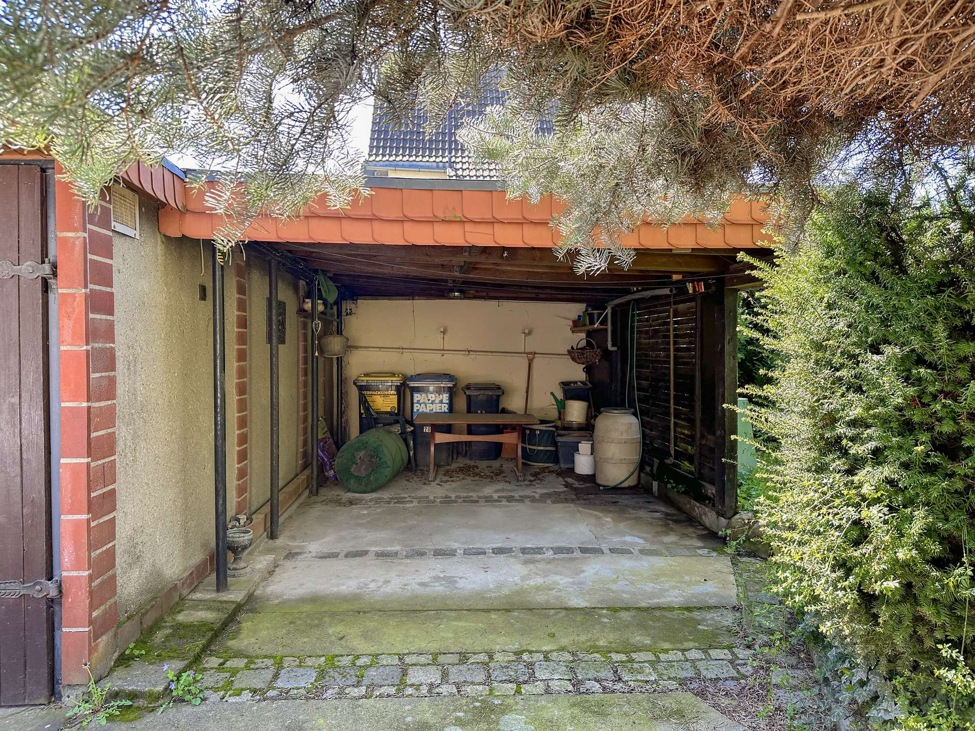 Open-front garage or carport with a tiled roof, cluttered with bins, a table, and gardening tools; overhanging branches frame the top and mossy steps lie at the entrance on the left and right side greenery.