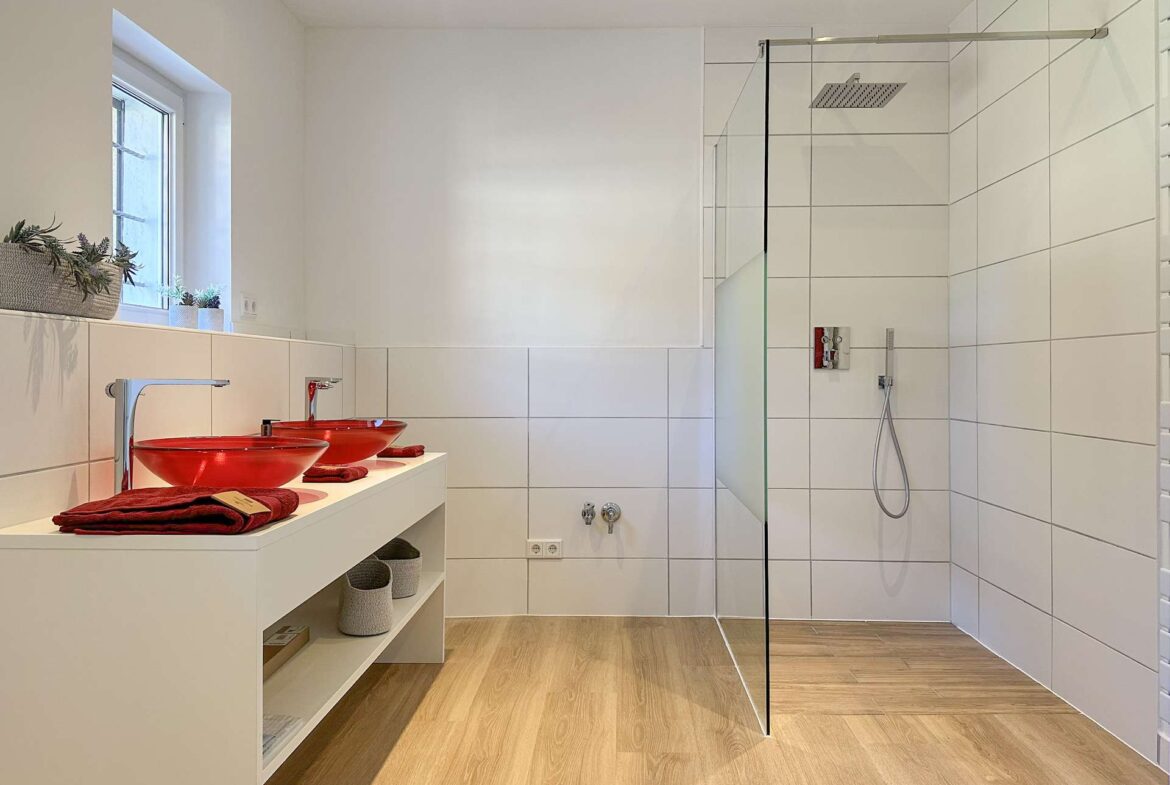 Modern bathroom with white tiles, wood-look floor, and a glass shower enclosure; red vessel sinks on a white vanity. Gently lit by a window.",